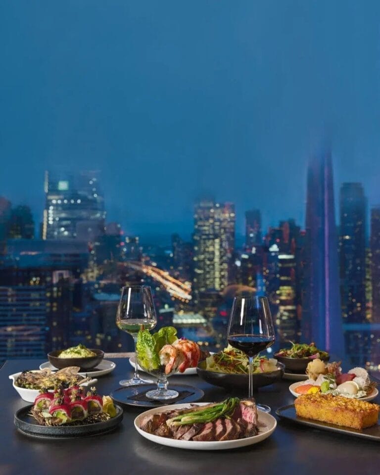 A table filled with gourmet dishes and wine glasses set against a glowing nighttime view of Toronto’s skyline, perfect for a fancy Father’s Day dinner near CN Tower.