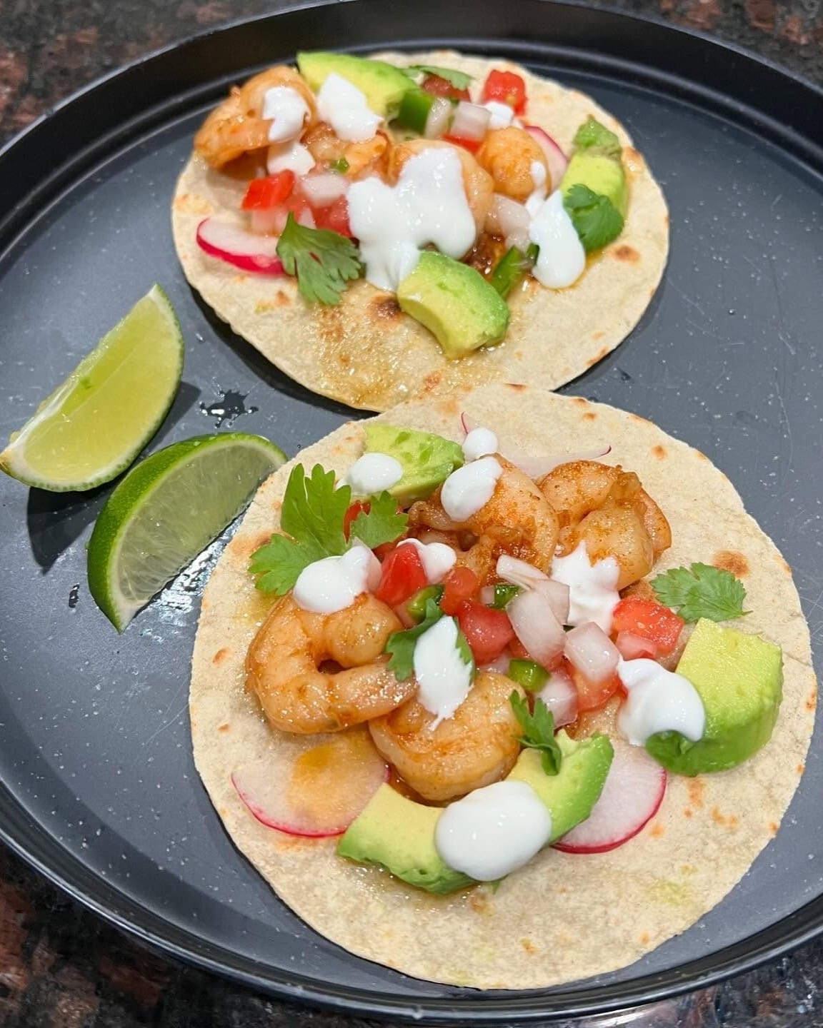 Shrimp tacos with spicy lime crema on corn tortillas, topped with diced tomatoes, radish slices, avocado, and fresh cilantro, served with lime wedges on a black plate.