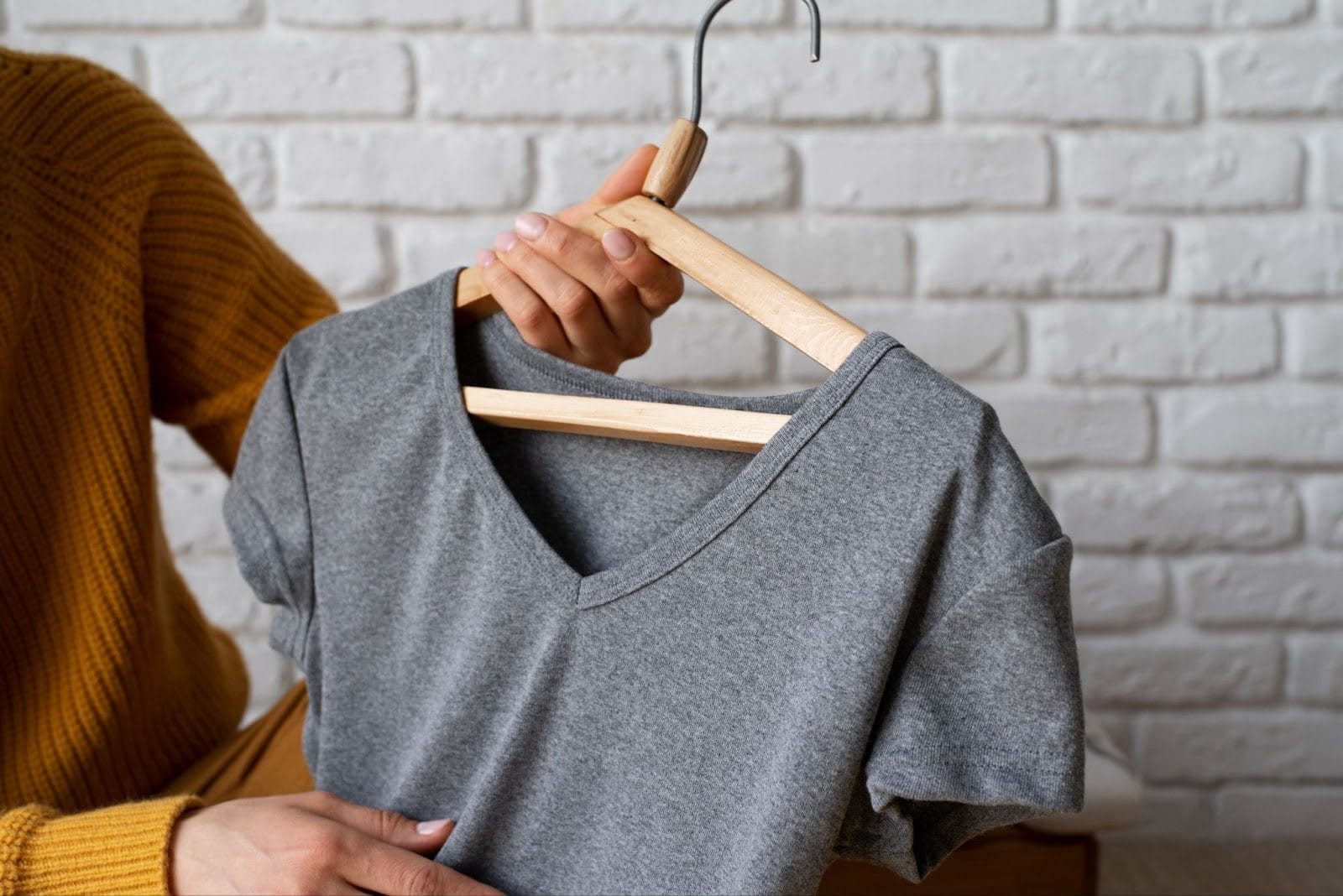 A person holding up a plain gray V-neck t-shirt on a wooden hanger, against a white brick wall.