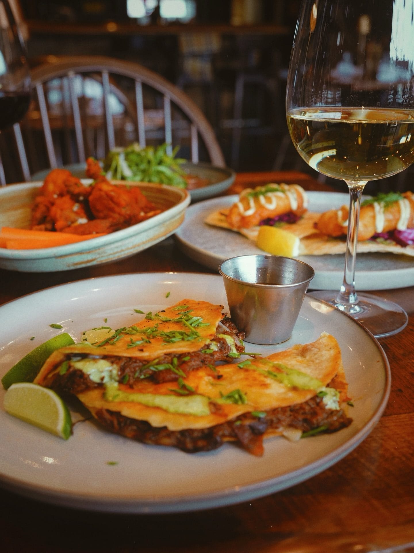 Kitchen on Sixth with beef quesadillas, lime wedges, fried bites, salad, and a glass of white wine on a wooden table.