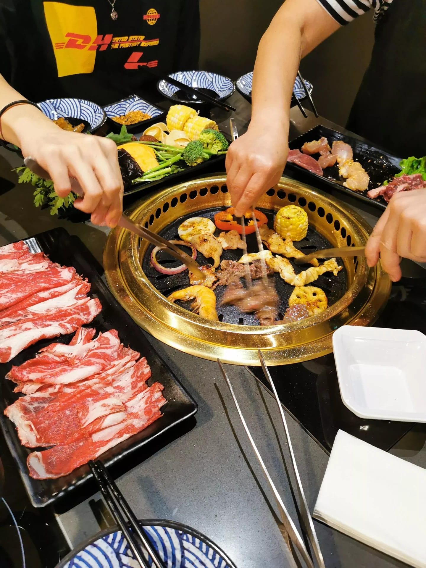 Diners grilling meat, seafood, and vegetables on a tabletop BBQ grill with sliced beef platters at Mix2 Grill & Hot Pot.